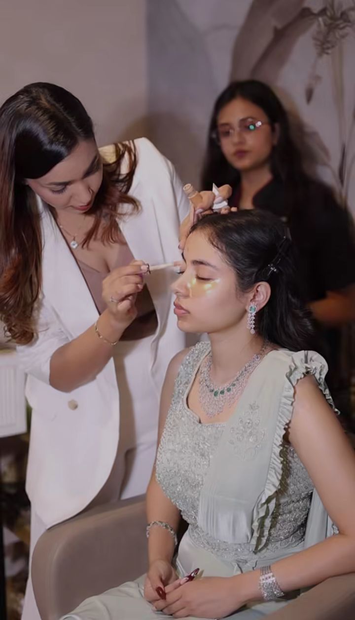 Makeup instructor applying concealer to a seated model while a student watches, in the Elaya School of Makeup studio