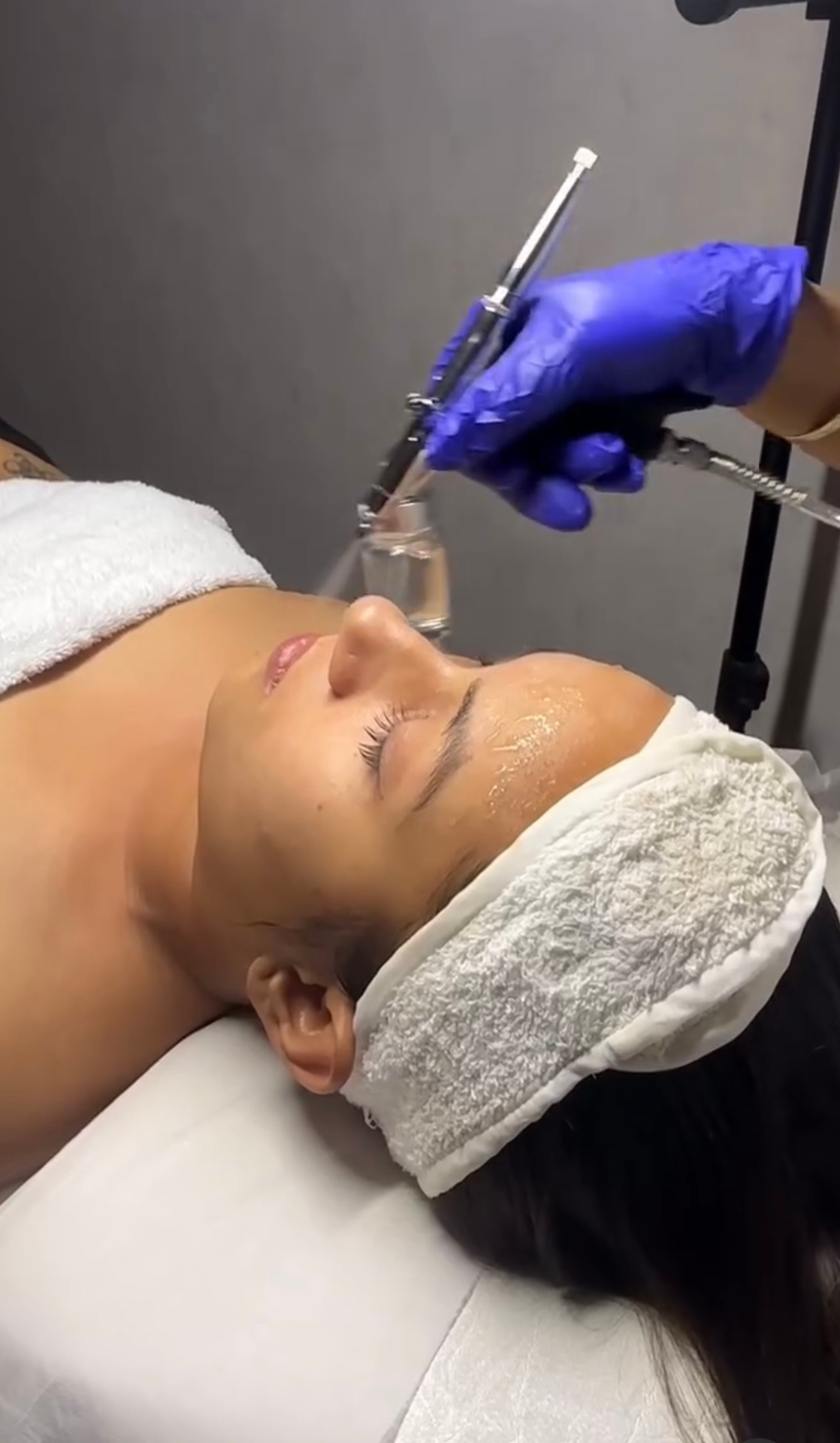 Client lying back with a white headband while a facial treatment is applied with a fine-mist spray device