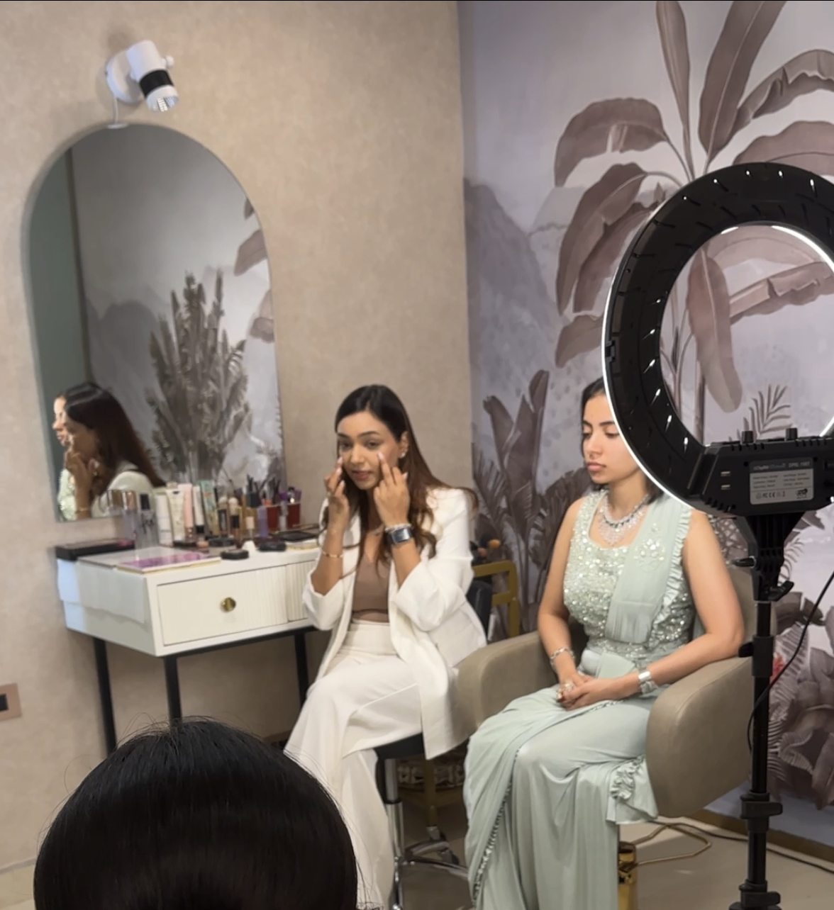 Makeup instructor demonstrating technique on a seated model beside a ring light while students watch, at Elaya School of Makeup